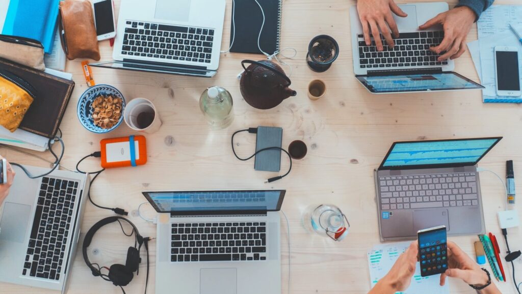 Busy collaboration space with multiple laptops, snacks, phones, and drinks