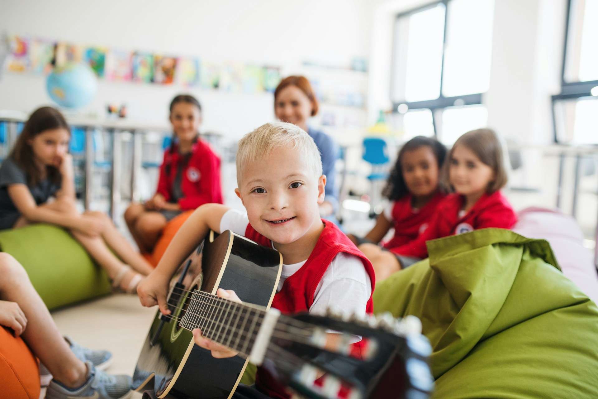 A happy young boy with Down syndrome plays a guitar in a classroom, surrounded by classmates. Reliable paging systems enhance communication and engagement in inclusive learning environments.