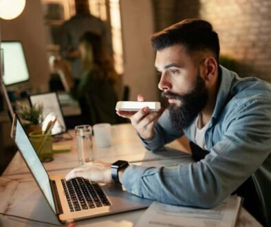 A bearded man sitting at a desk in a modern office, speaking into his smartphone with a laptop open in front of him, demonstrating the use of voice recognition technology for business tasks.