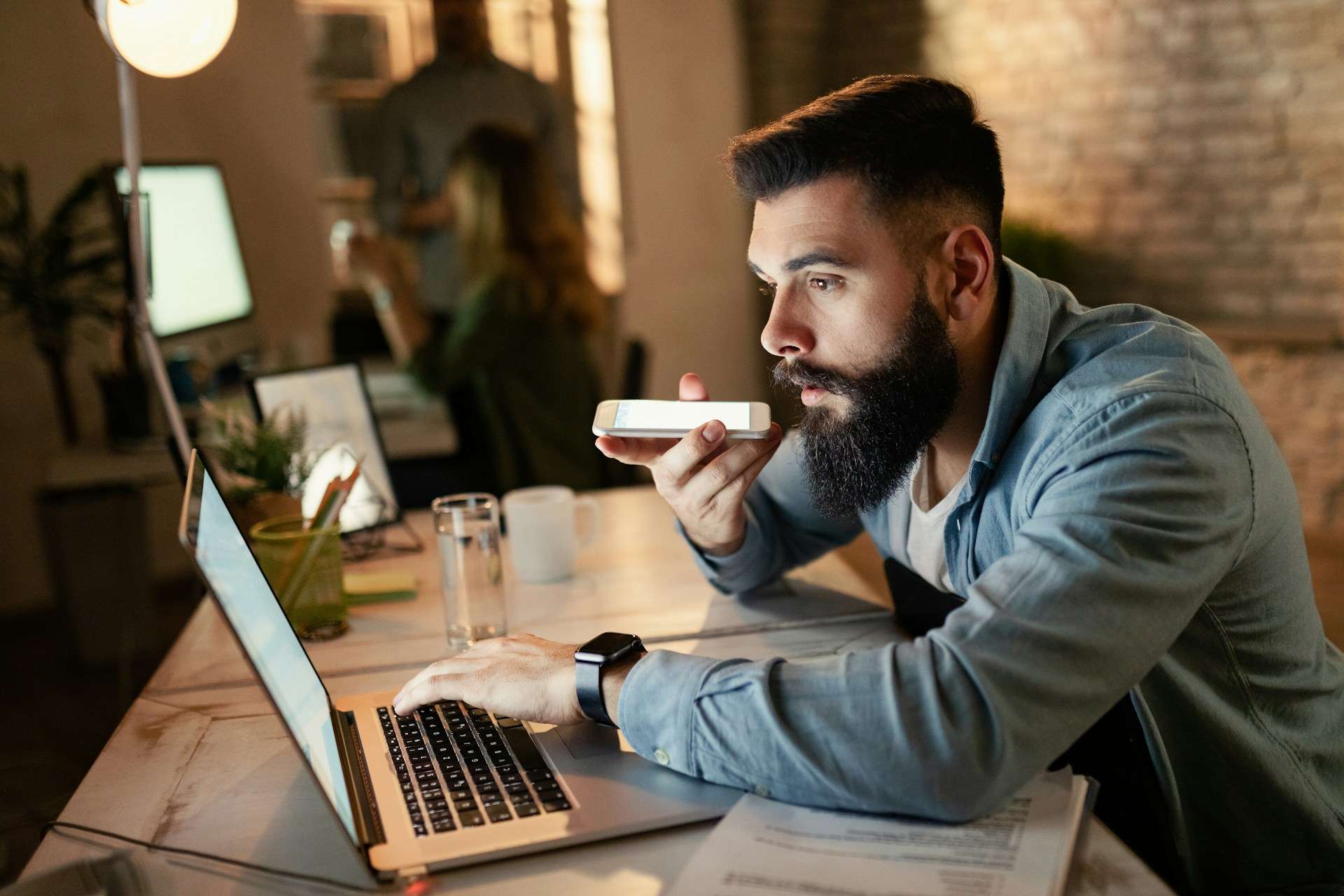 A bearded man sitting at a desk in a modern office, speaking into his smartphone with a laptop open in front of him, demonstrating the use of voice recognition technology for business tasks.