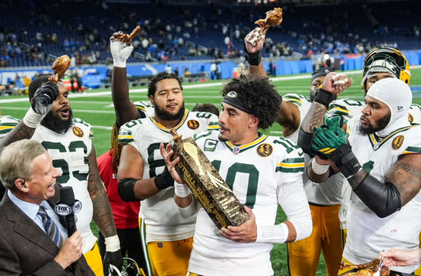 Green Bay Packers players celebrating on the field after a game, holding turkey legs and a trophy during a post-game interview.